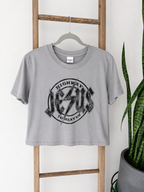 A light gray cropped t-shirt with a distressed black graphic reading 'JESUS HIGHWAY TO HEAVEN' hanging on a wooden hanger from a wooden ladder, with a snake plant in a pot on the right.