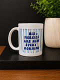 A white ceramic coffee mug with blue text reading 'HIS MERCIES ARE NEW EVERY MORNING' and blue vertical stripe accents, displayed on a black tray with a potted plant in the background.