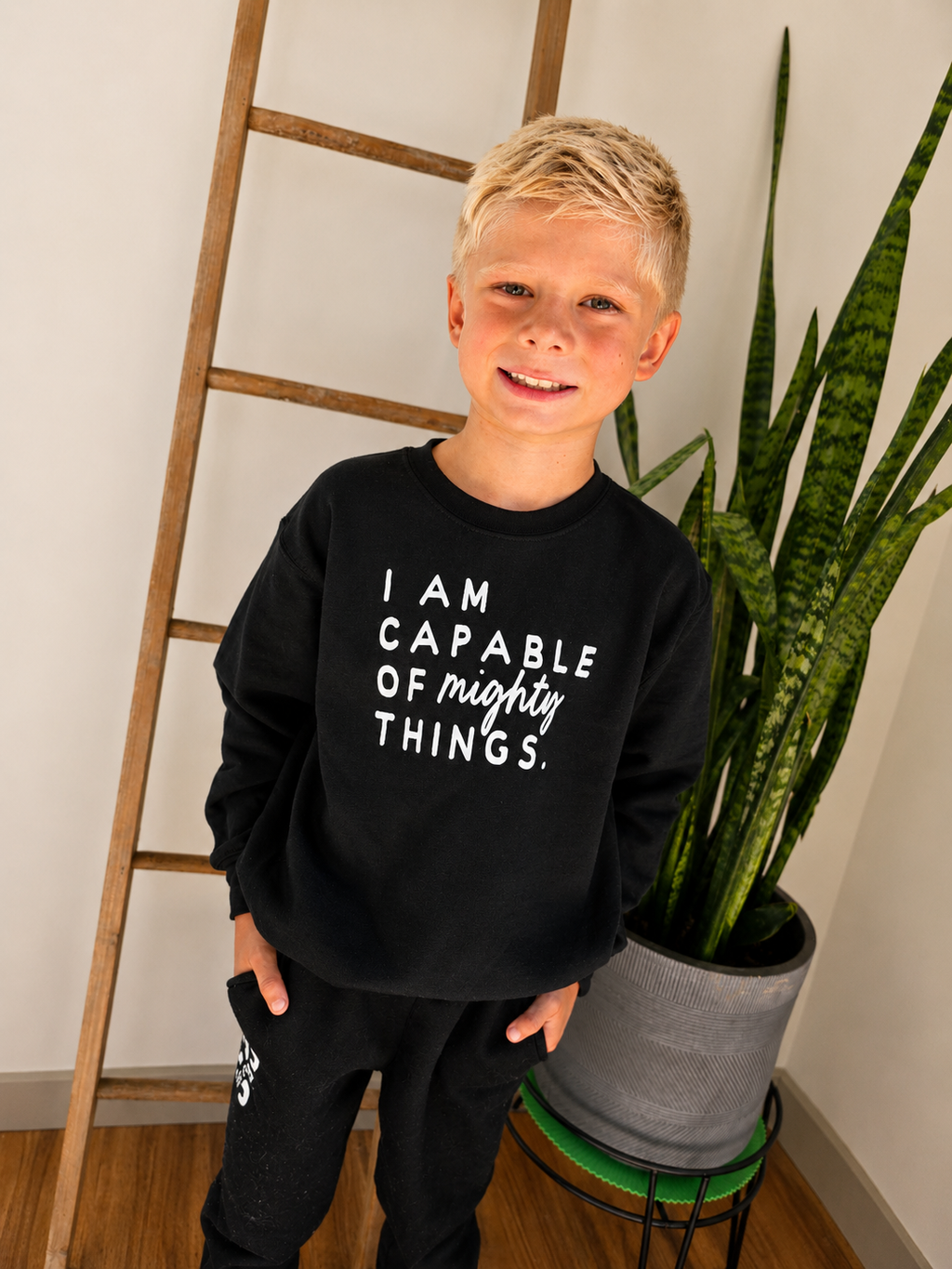 A young boy with blonde hair stands indoors wearing a black sweatshirt with the white printed text 'I AM CAPABLE OF mighty THINGS.' and black sweatpants.