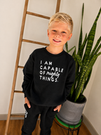A young boy with blonde hair stands indoors wearing a black sweatshirt with the white printed text 'I AM CAPABLE OF mighty THINGS.' and black sweatpants.