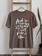 A brown short-sleeve t-shirt with white script text reading 'And in every season I will praise Him' hanging on a wooden ladder against a light wall, with a plant visible on the right.