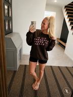 A woman takes a mirror selfie in a home hallway wearing a dark brown crewneck sweatshirt with pink text that reads 'IN HIS PRESENCE THERE IS PEACE.' She is also wearing black shorts.