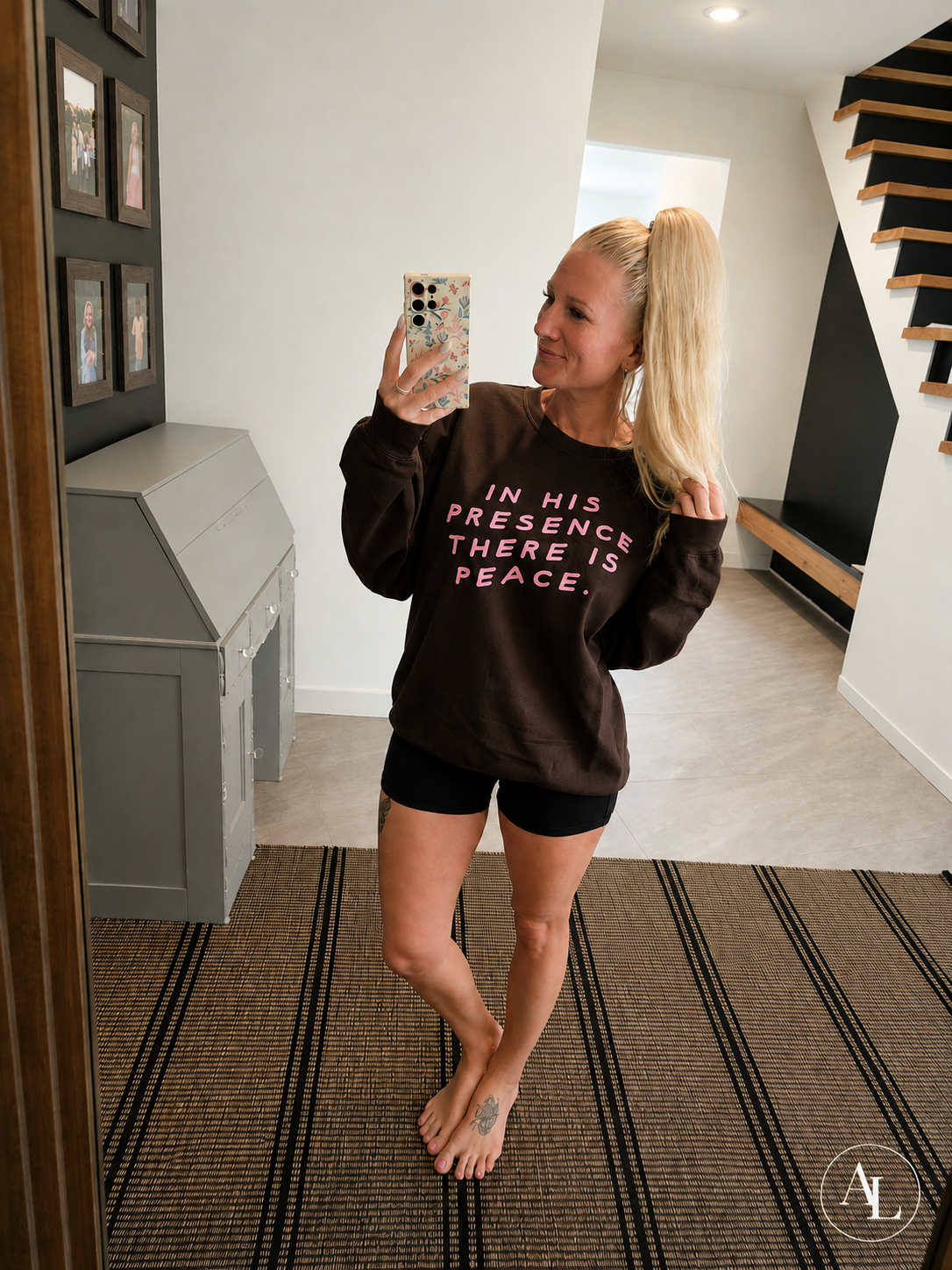 A woman takes a mirror selfie in a home hallway wearing a dark brown crewneck sweatshirt with pink text that reads 'IN HIS PRESENCE THERE IS PEACE.' She is also wearing black shorts.
