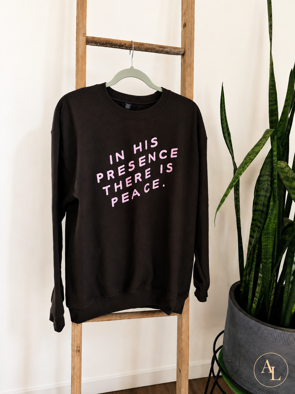 A brown crewneck sweatshirt with the text 'IN HIS PRESENCE THERE IS PEACE.' in light pink, hanging on a wooden ladder next to a green snake plant.