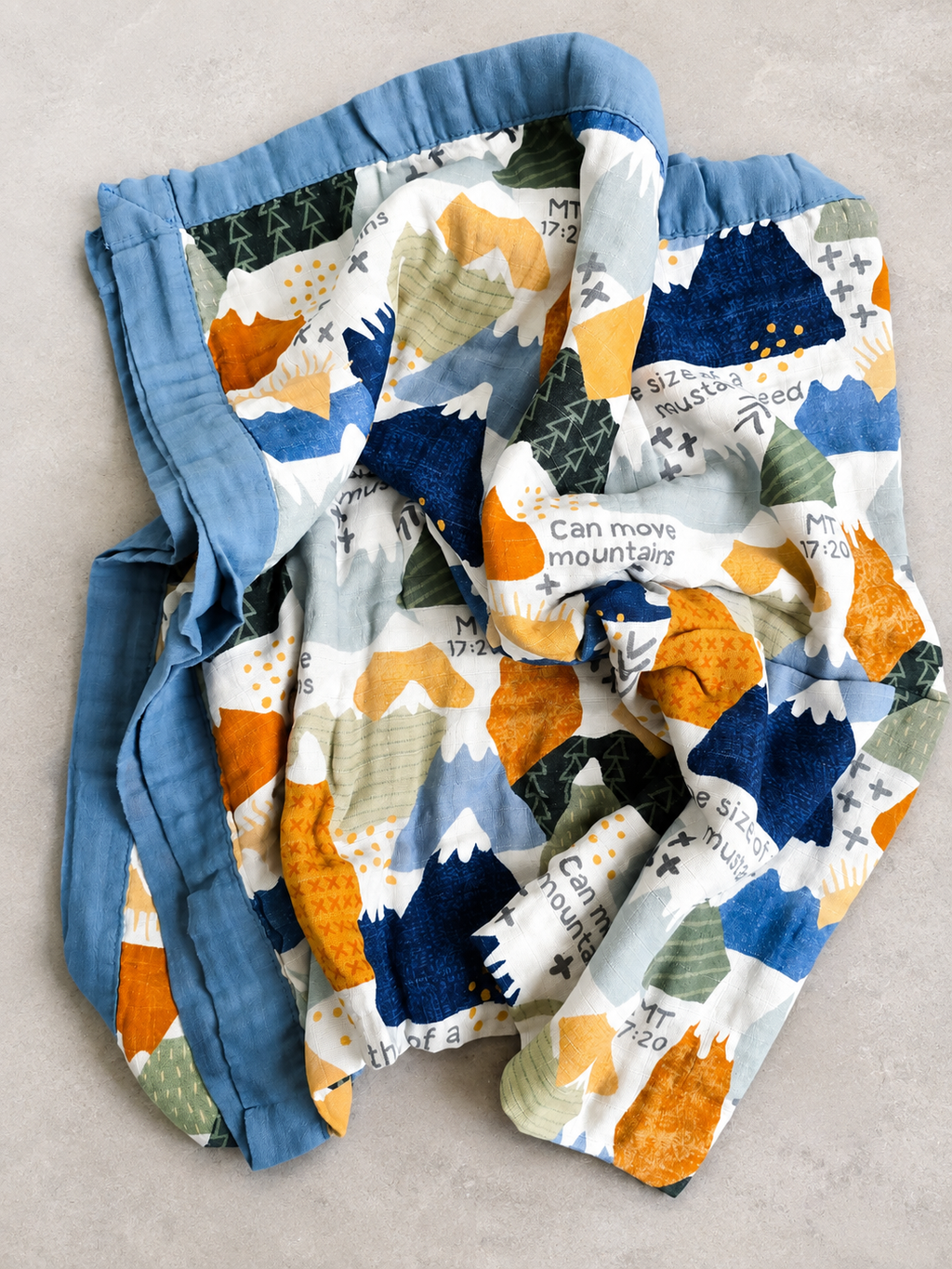 A folded quilted baby blanket with a colorful mountain and fox pattern, featuring a solid blue ruffled border, laid flat on a light gray textured surface.
