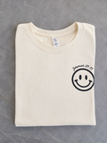 A folded cream-colored t-shirt with a black smiley face graphic and the text 'Jeremiah 29:11' printed on the left chest, laid flat on a gray tiled surface.