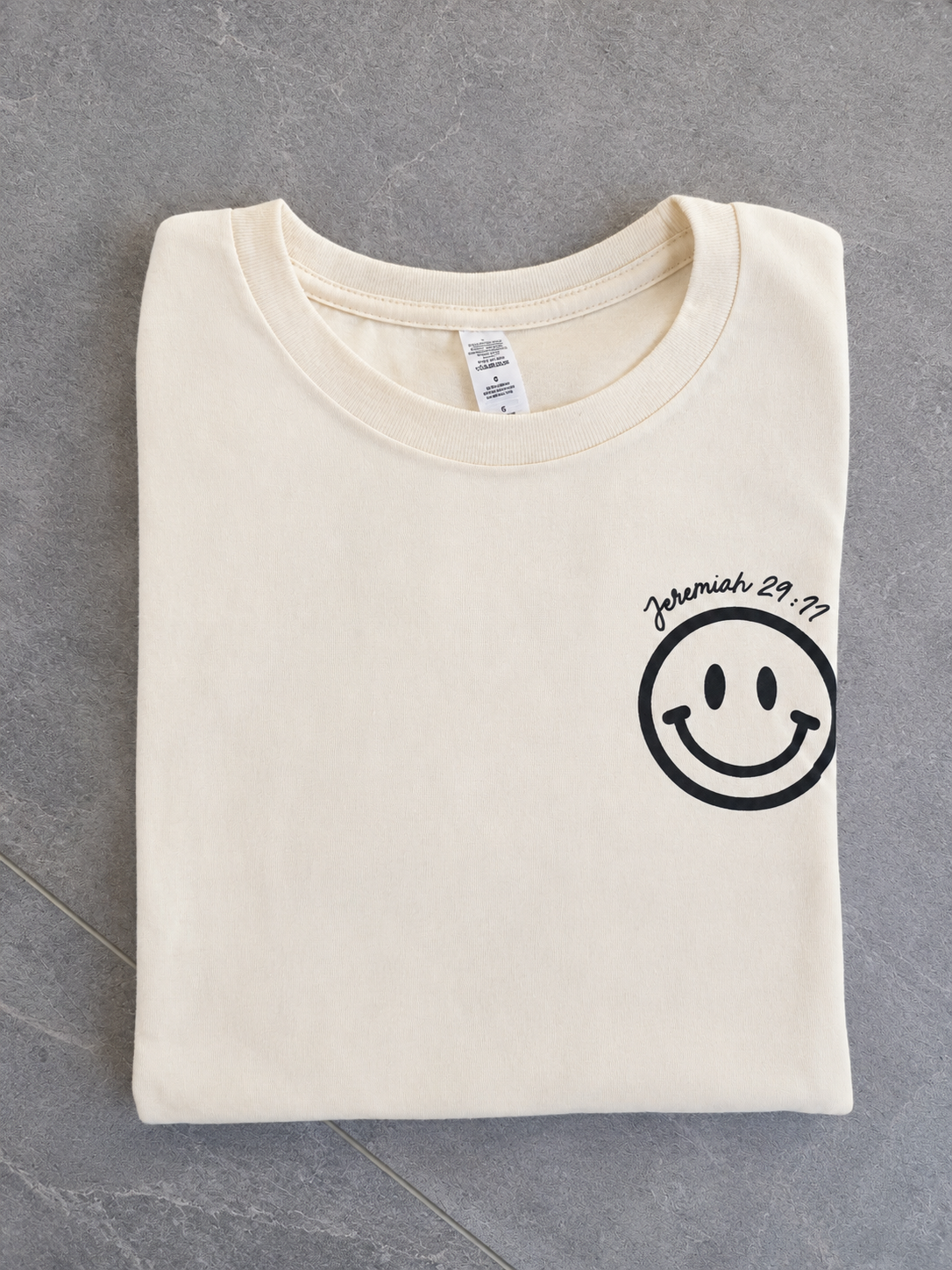 A folded cream-colored t-shirt with a black smiley face graphic and the text 'Jeremiah 29:11' printed on the left chest, laid flat on a gray tiled surface.