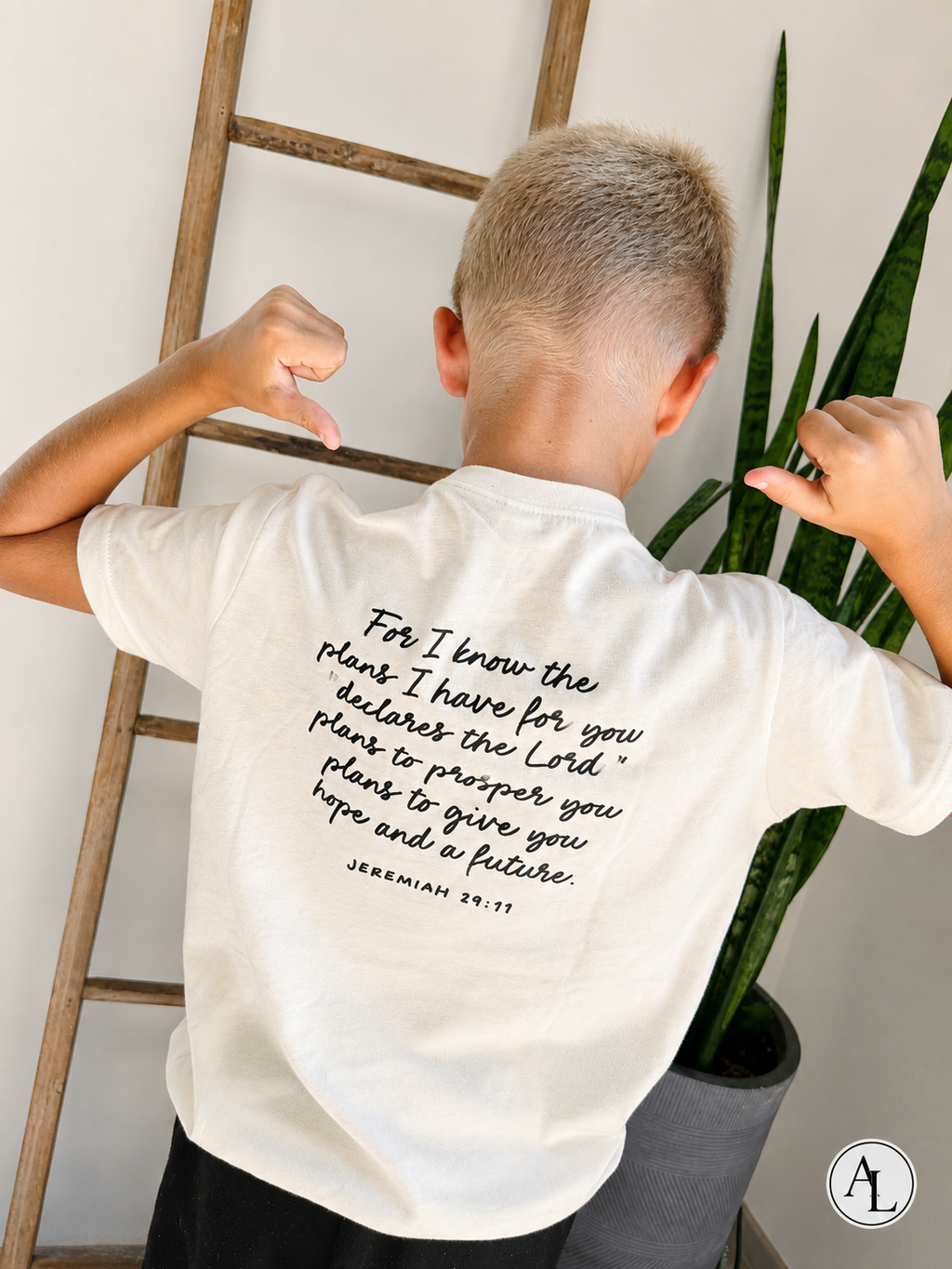 A child viewed from behind wearing a white t-shirt with a black printed Bible verse from Jeremiah 29:11. The child is posing with hands on hips in front of a wooden ladder and a green snake plant.
