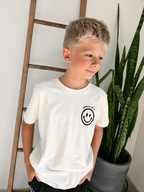 A young boy wearing a white short-sleeve t-shirt with a black smiley face graphic on the left chest, standing indoors next to a wooden ladder and a potted snake plant.