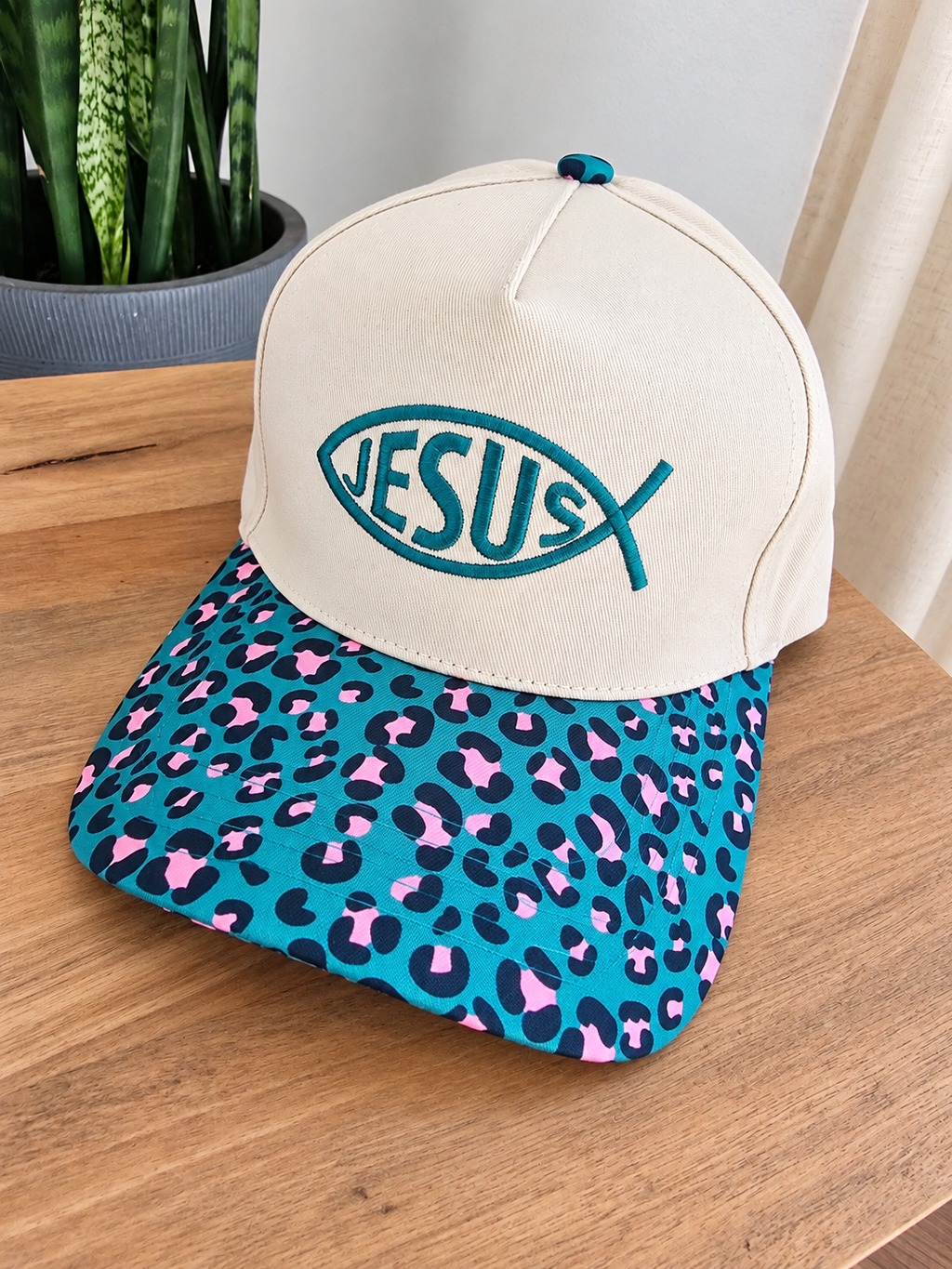 A baseball cap resting on a wooden table, featuring an off-white crown with a teal embroidered fish-shaped Jesus logo, and a teal brim with a pink and black leopard print.