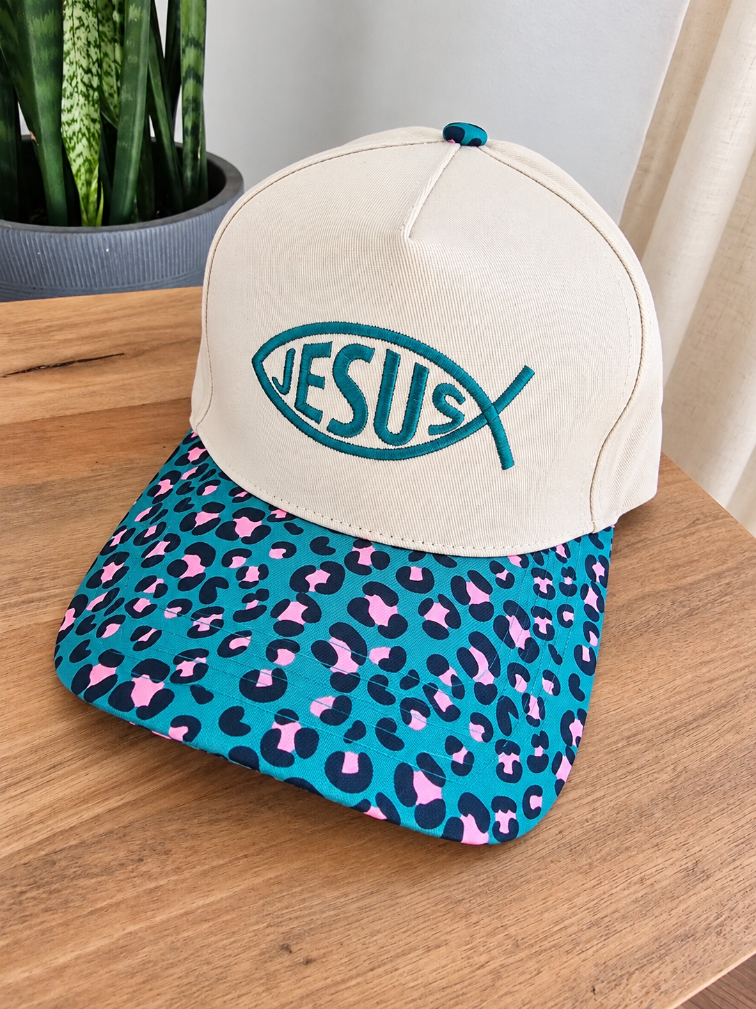 A baseball cap resting on a wooden table, featuring an off-white crown with a teal embroidered fish-shaped Jesus logo, and a teal brim with a pink and black leopard print.