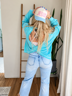 A woman is shown from behind wearing a light blue long-sleeve crop top, light-wash flare jeans, and a light blue baseball cap with 'JESUS' written in pink on the front. She is posing in a room with a wooden ladder, plants, and a window.
