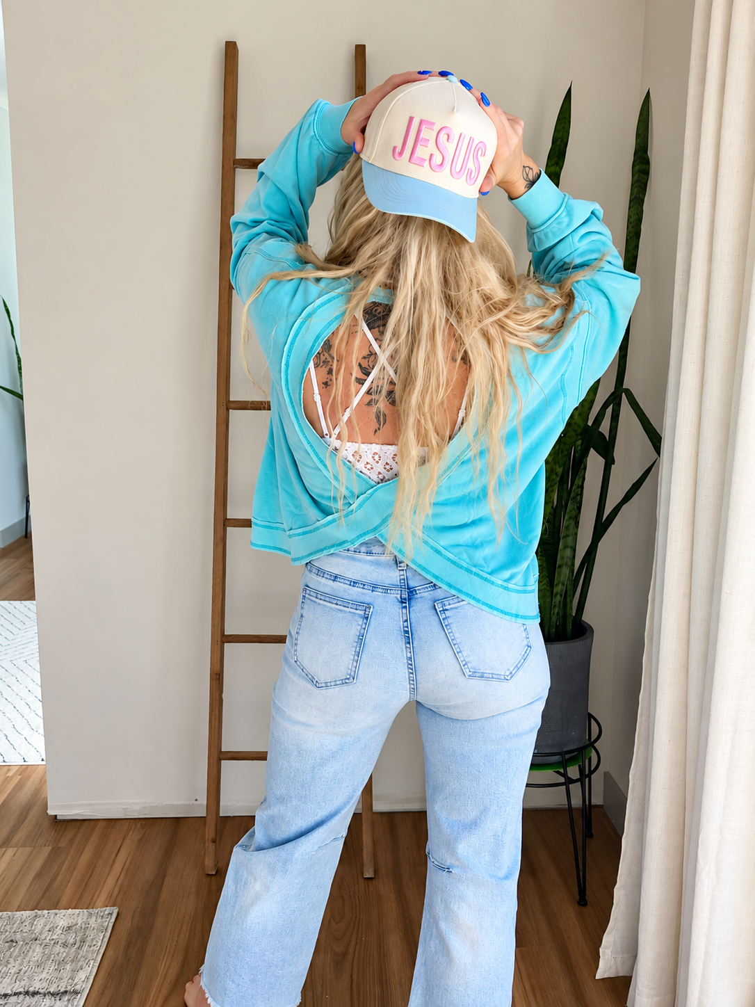 A woman is shown from behind wearing a light blue long-sleeve crop top, light-wash flare jeans, and a light blue baseball cap with 'JESUS' written in pink on the front. She is posing in a room with a wooden ladder, plants, and a window.