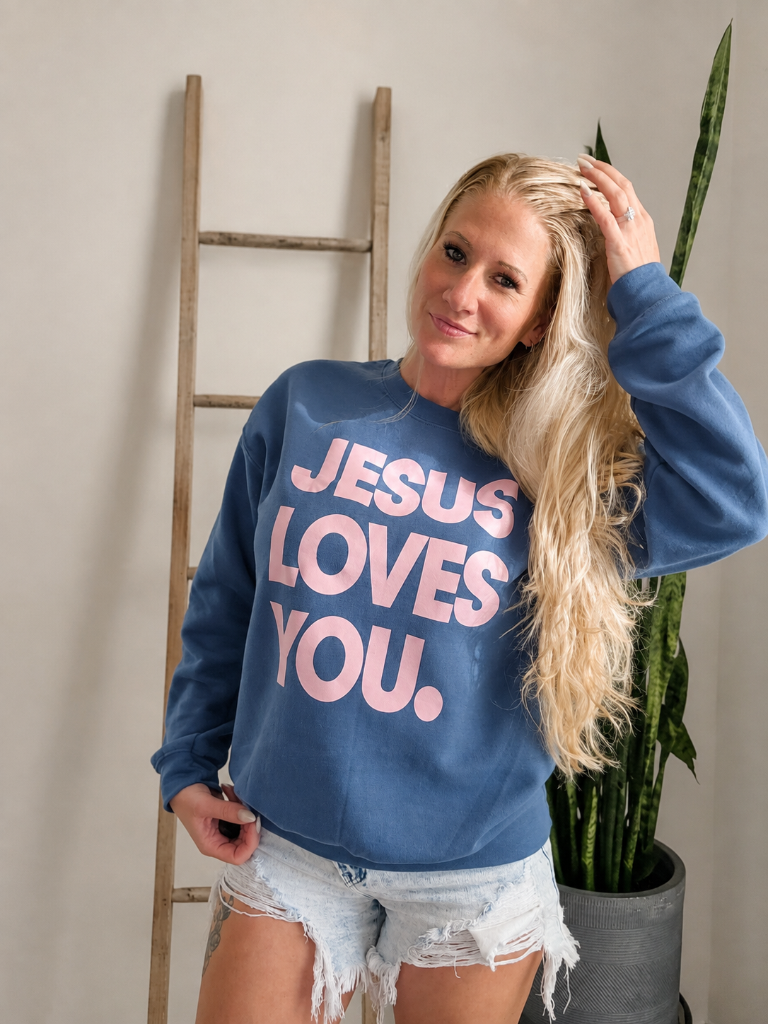 A blonde woman wearing a blue crewneck sweatshirt with the text 'JESUS LOVES YOU.' in pink, paired with light-wash distressed denim shorts, standing in a room with a wooden ladder and a potted snake plant.