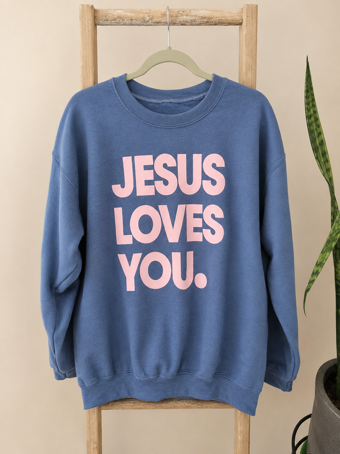 A blue crewneck sweatshirt hanging on a wooden hanger from a wooden ladder, featuring the text 'JESUS LOVES YOU.' in light pink.