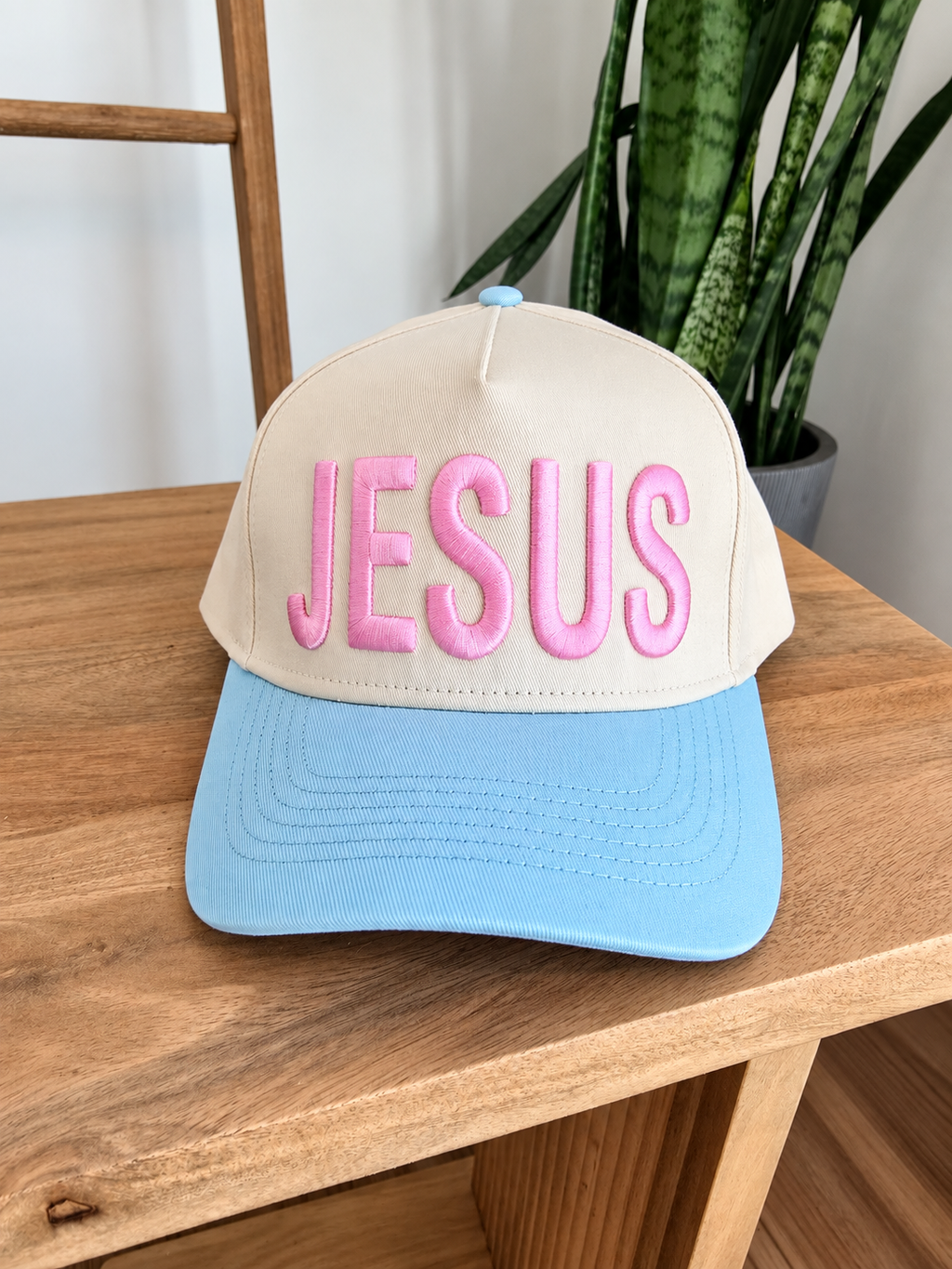 A beige and light blue trucker-style cap with the word 'JESUS' embroidered in pink across the front, displayed on a wooden table with a plant in the background.
