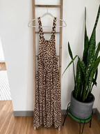 A sleeveless leopard-print maxi dress with wide shoulder straps hangs on a wooden ladder indoors, next to a potted snake plant.