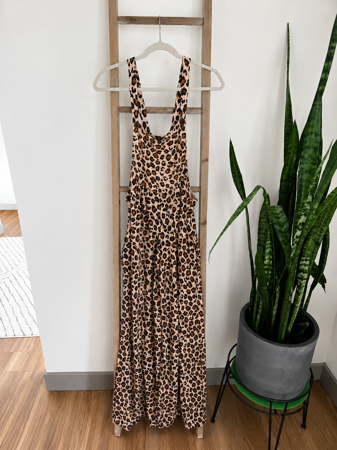 A sleeveless leopard-print maxi dress with wide shoulder straps hangs on a wooden ladder indoors, next to a potted snake plant.