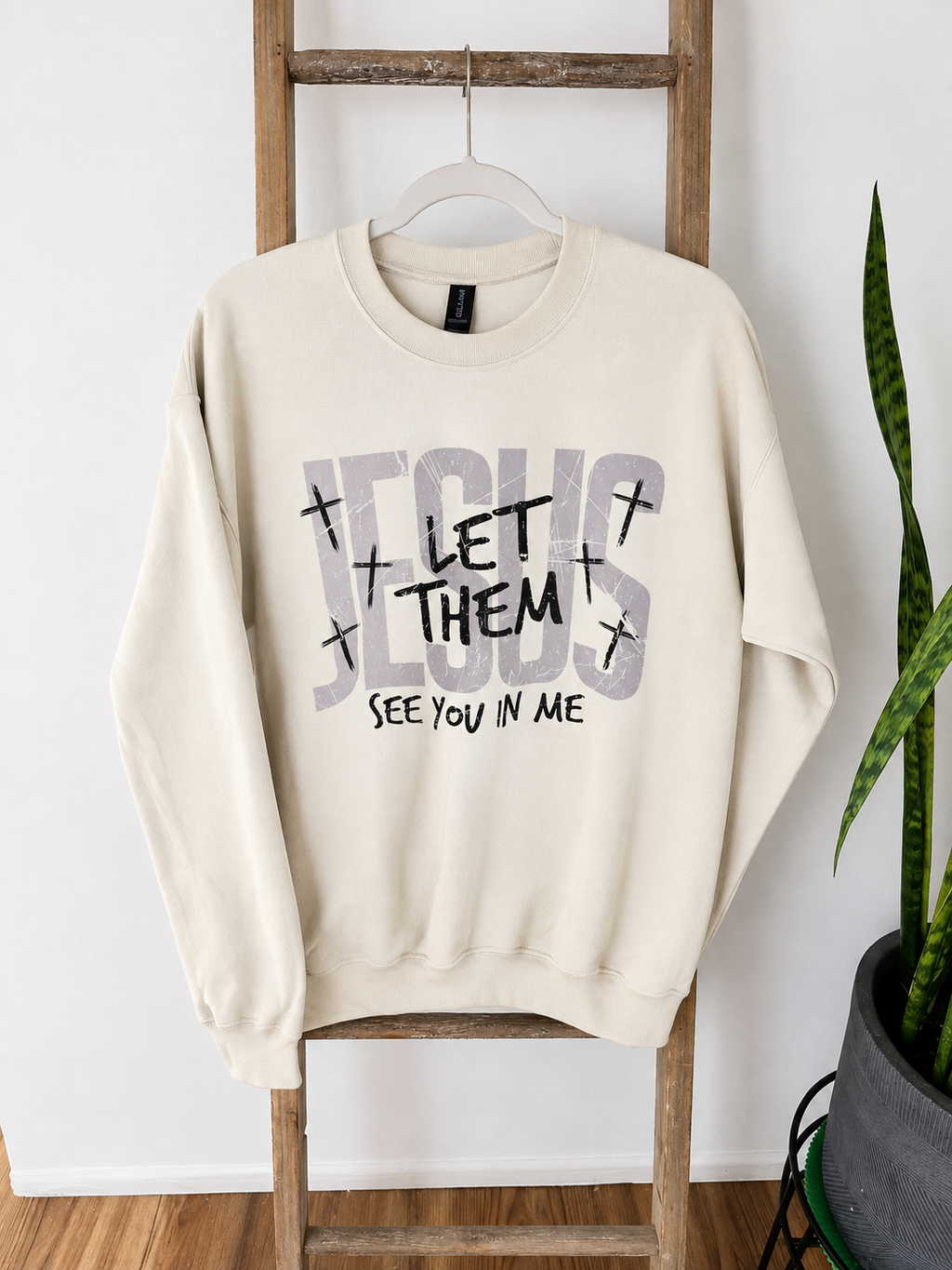 An off-white crewneck sweatshirt hanging on a wooden ladder, featuring a graphic print that reads 'JESUS LET THEM SEE YOU IN ME' with small crosses.