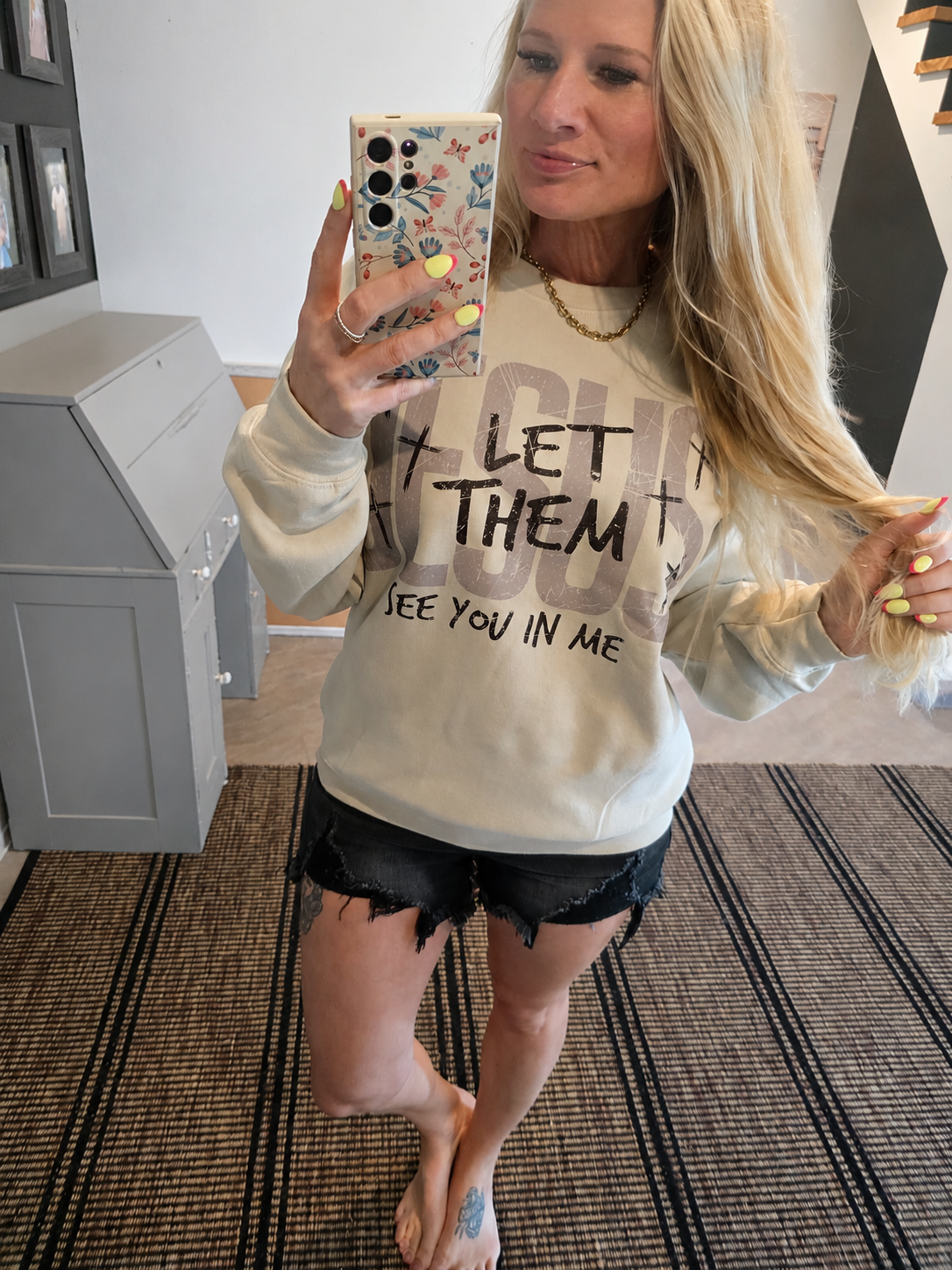 A woman taking a mirror selfie in an indoor room, wearing a cream-colored sweatshirt with a graphic that reads 'LET THEM SEE YOU IN ME' and black frayed denim shorts.