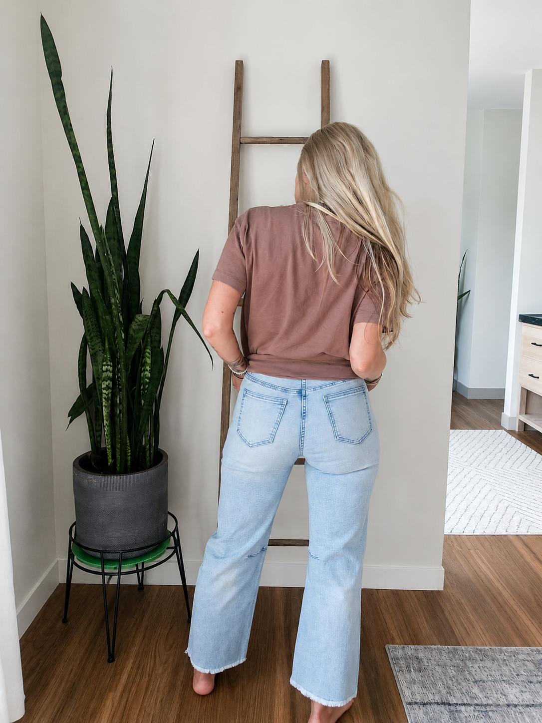 A woman stands with her back to the camera in a bright room, wearing a brown short-sleeve t-shirt and light-wash blue flare jeans. She is positioned next to a large potted snake plant and a wooden ladder against a white wall.