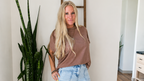 A woman with long blonde hair stands barefoot in a home interior, wearing a loose taupe-brown short-sleeve t-shirt and light-wash blue cropped jeans. She is posing next to a wooden ladder shelf, a large potted snake plant on a black stand, and a rug on a hardwood floor.