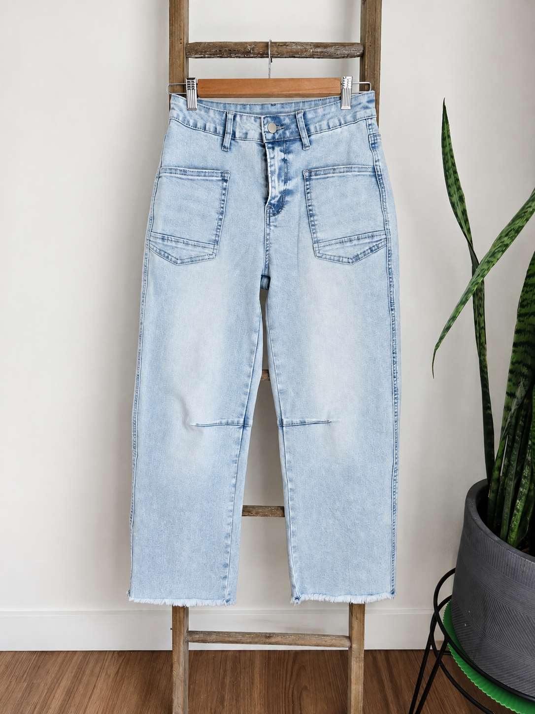 Light wash blue denim jeans with a straight, slightly cropped leg and raw frayed hems, hanging on a wooden hanger on a rustic wooden ladder next to a potted snake plant.