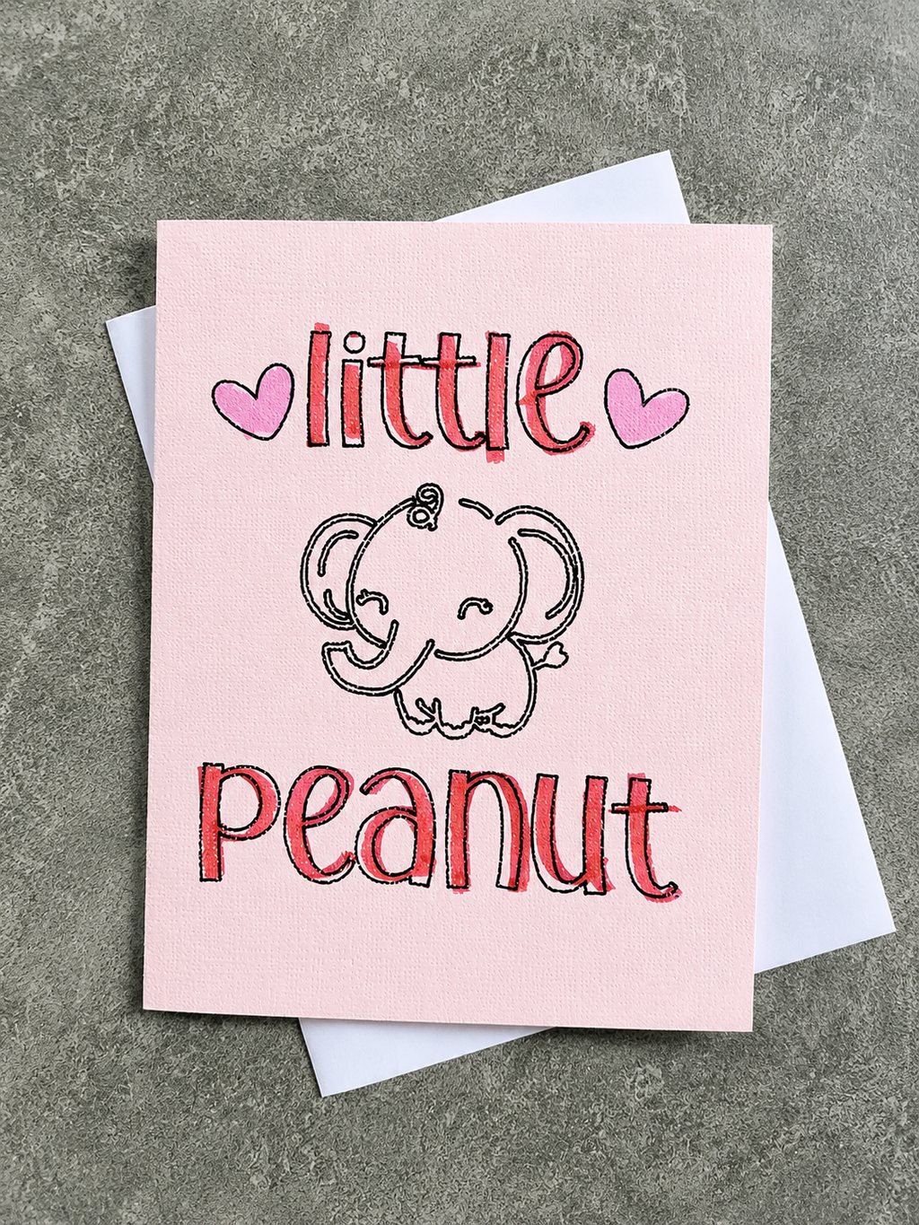 A pink greeting card with a cute cartoon elephant and the words 'little peanut' in red outlined lettering, shown with a white envelope on a textured gray surface.