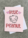 A pink greeting card with a cute cartoon elephant and the words 'little peanut' in red outlined lettering, shown with a white envelope on a textured gray surface.