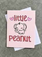 A pink greeting card with a cute cartoon elephant and the words 'little peanut' in red outlined lettering, shown with a white envelope on a textured gray surface.