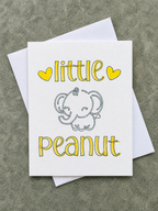 A white greeting card with the words 'little peanut' in yellow hand-lettered style text and a simple gray elephant illustration, shown with a white envelope on a textured gray surface.