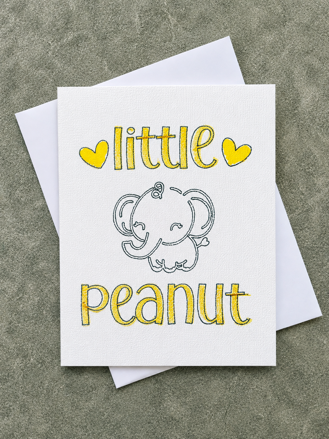 A white greeting card with the words 'little peanut' in yellow hand-lettered style text and a simple gray elephant illustration, shown with a white envelope on a textured gray surface.