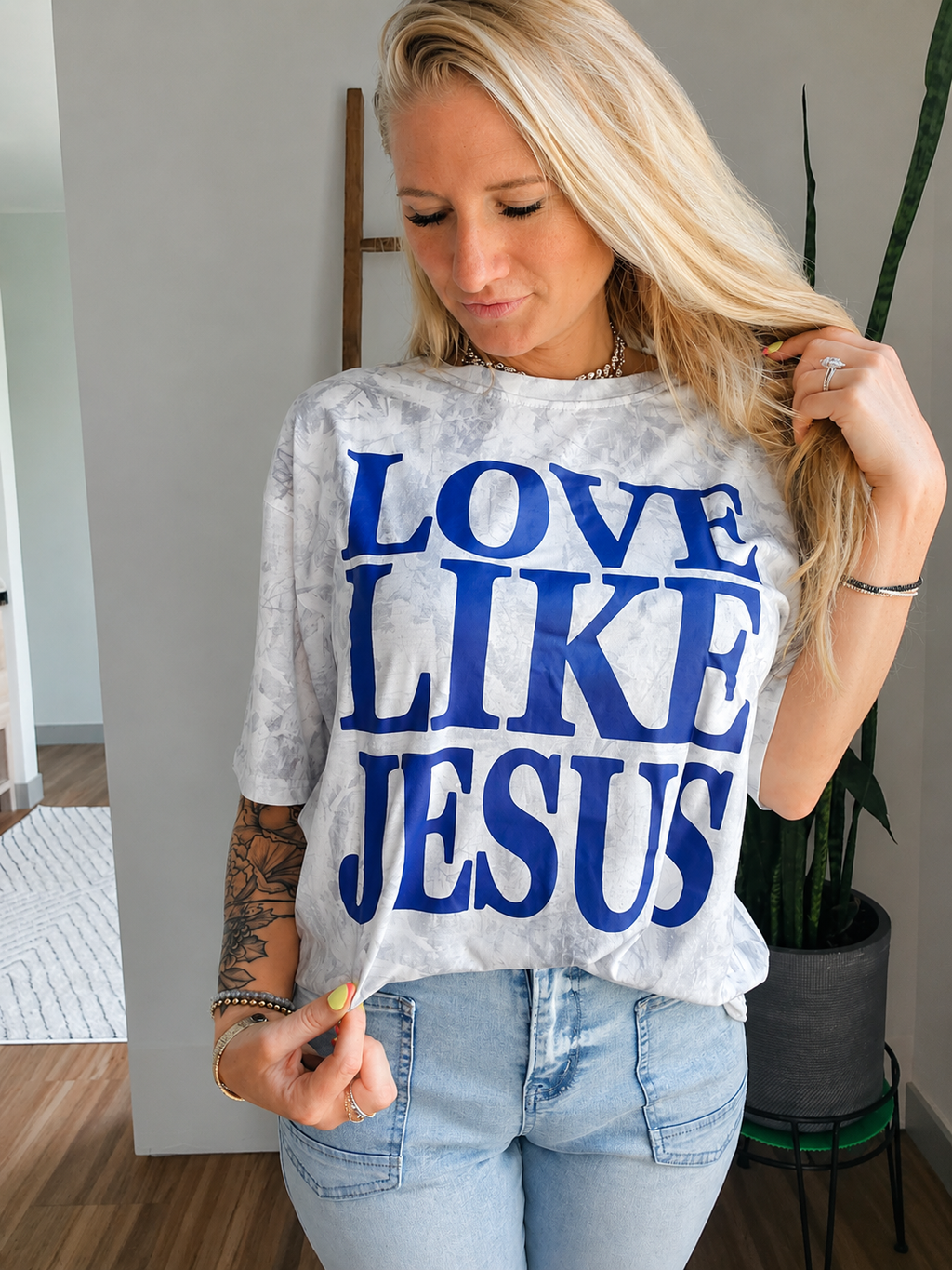 A woman wearing a white and gray tie-dye t-shirt with the text 'LOVE LIKE JESUS' printed in bold blue letters, paired with light-wash blue jeans.
