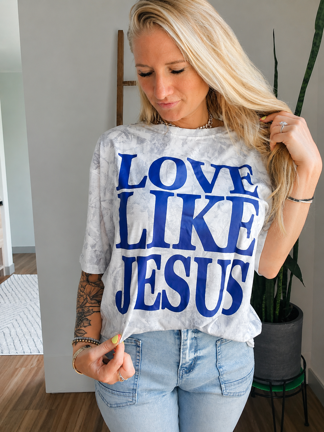 A woman wearing a white and gray tie-dye t-shirt with the text 'LOVE LIKE JESUS' printed in bold blue letters, paired with light-wash blue jeans.