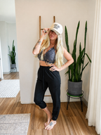 A woman stands barefoot in a bright indoor room wearing a white baseball cap with leopard print trim and the text 'SWEAT LIKE A GIRL', a gray lace sports bra, and black cropped sweatpants.