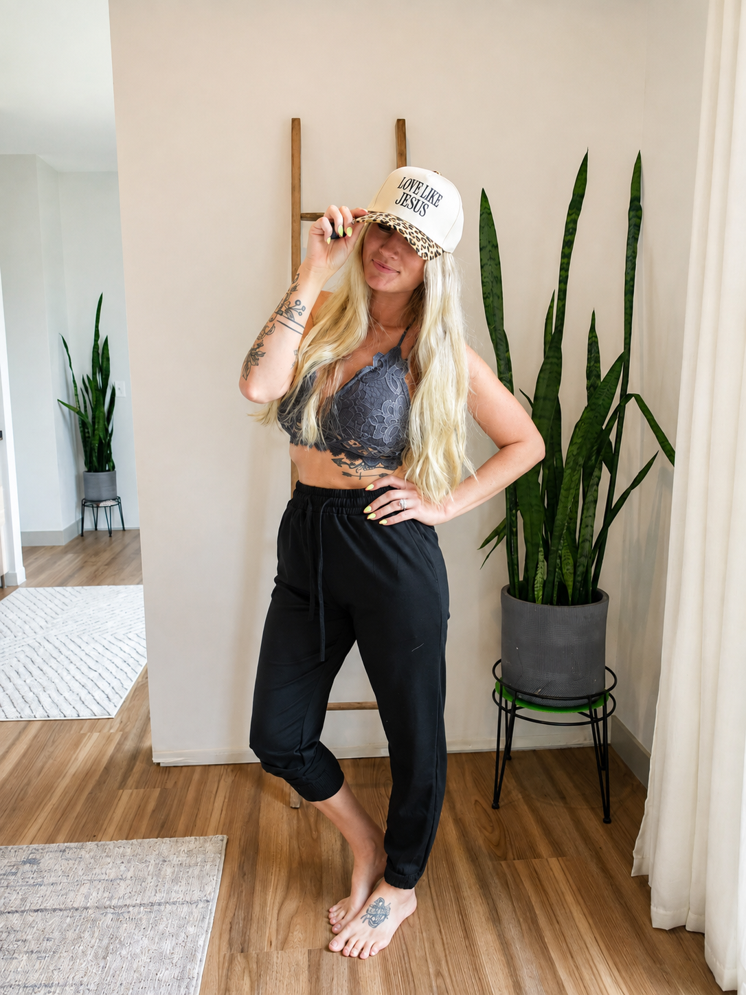 A woman stands barefoot in a bright indoor room wearing a white baseball cap with leopard print trim and the text 'SWEAT LIKE A GIRL', a gray lace sports bra, and black cropped sweatpants.