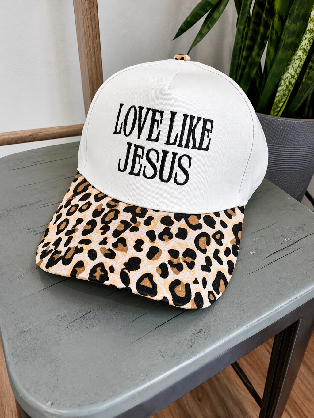 A baseball cap with a white front panel and black text reading 'LOVE LIKE JESUS' sits on a gray table. The brim has a tan, brown, and black leopard print pattern.
