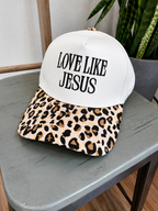 A baseball cap with a white front panel and black text reading 'LOVE LIKE JESUS' sits on a gray table. The brim has a tan, brown, and black leopard print pattern.