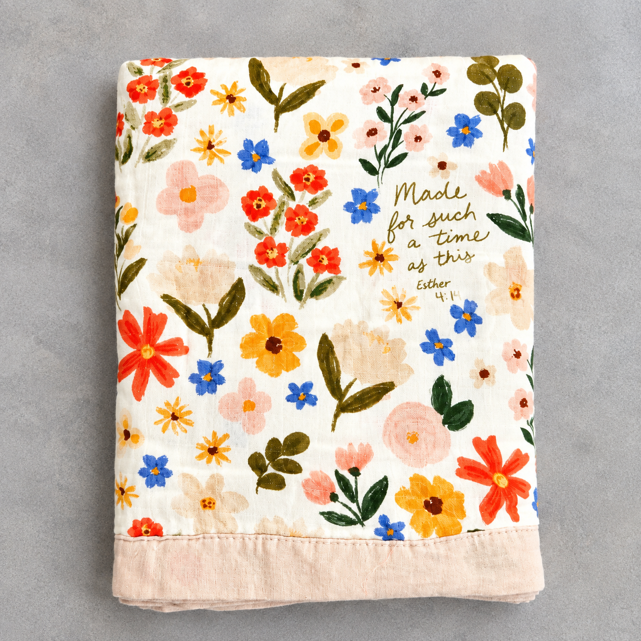 A folded cotton blanket with a cream background, featuring a colorful embroidered floral pattern in red, orange, yellow, blue, pink, and green, with a peach-colored ribbed hem and gold cursive text reading 'Made for such a time as this Esther 1:10'.