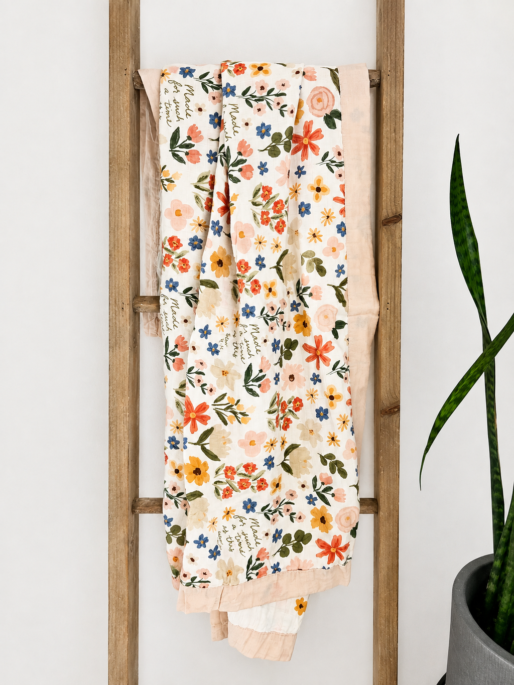 A cream-colored cotton blanket with a colorful floral and script pattern, draped over a rustic wooden ladder next to a potted snake plant.
