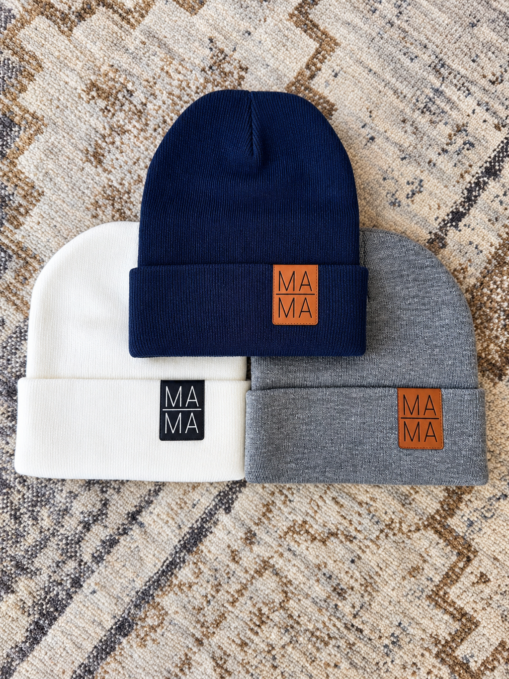 Three knit beanie hats arranged in a flat lay on a patterned rug: one navy blue, one white, and one heather gray. Each hat has a folded cuff with a small square logo patch reading MAMA.