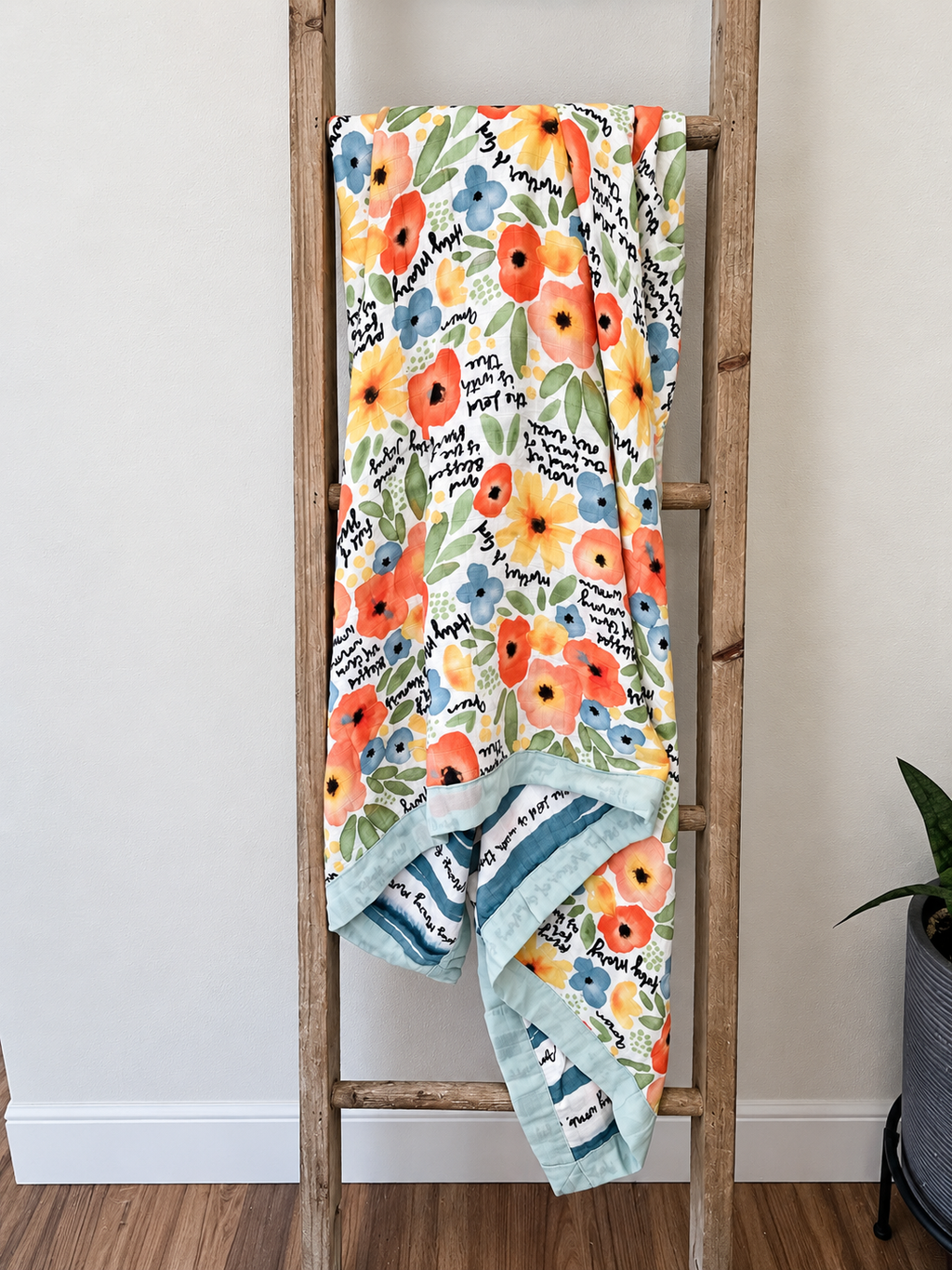 A floral baby blanket with orange, blue, and yellow flowers, green leaves, and black script text, draped over a rustic wooden ladder in a home setting.