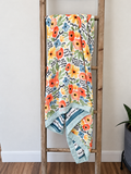A floral baby blanket with orange, blue, and yellow flowers, green leaves, and black script text, draped over a rustic wooden ladder in a home setting.