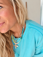 A close-up lifestyle shot of a woman wearing a layered necklace set. The necklace features a silver chain with a small cross pendant, layered with a strand of light blue beads and a separate strand of yellow and light blue beads.