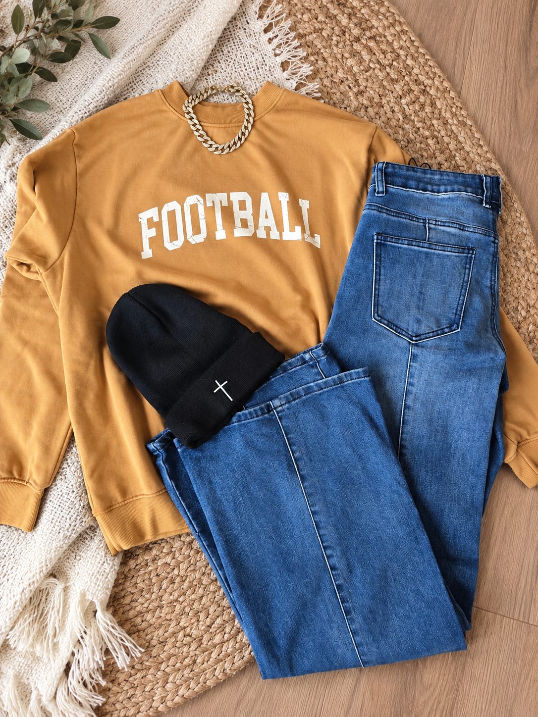 A styled flat-lay outfit featuring a mustard yellow sweatshirt with 'FOOTBALL' printed in white across the chest, paired with blue jeans, a black beanie with a small white cross, and a gold chain necklace, arranged on a woven rug over a cream knit blanket.