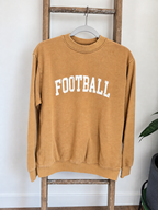 A washed tan crewneck sweatshirt with 'FOOTBALL' printed in white arched block letters across the chest, hanging on a hanger from a rustic wooden ladder against a white wall.