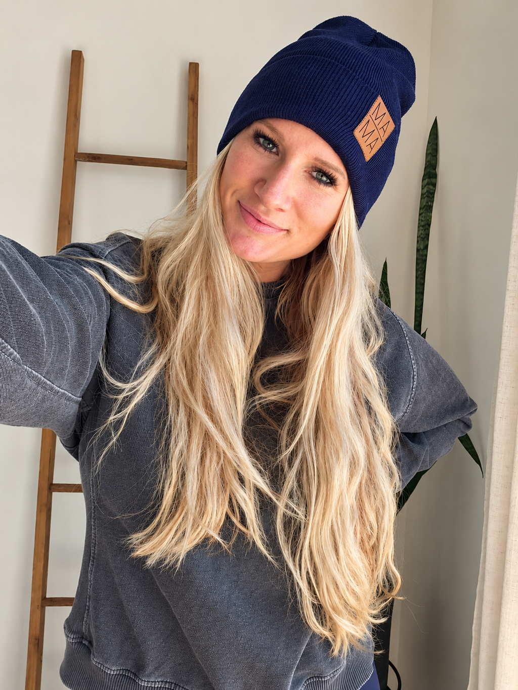 A woman with long blonde hair wearing a navy blue ribbed knit beanie with a tan patch on the side, paired with a dark gray sweatshirt in an indoor setting.