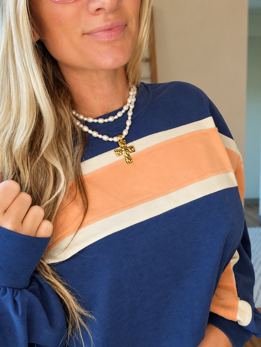 A close-up of a woman wearing a long-sleeve crewneck sweatshirt with horizontal stripes in navy blue, peach, and cream. She is also wearing a double-strand pearl necklace with a gold cross pendant.