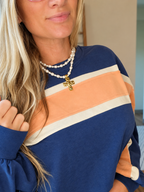A close-up of a woman wearing a long-sleeve crewneck sweatshirt with horizontal stripes in navy blue, peach, and cream. She is also wearing a double-strand pearl necklace with a gold cross pendant.