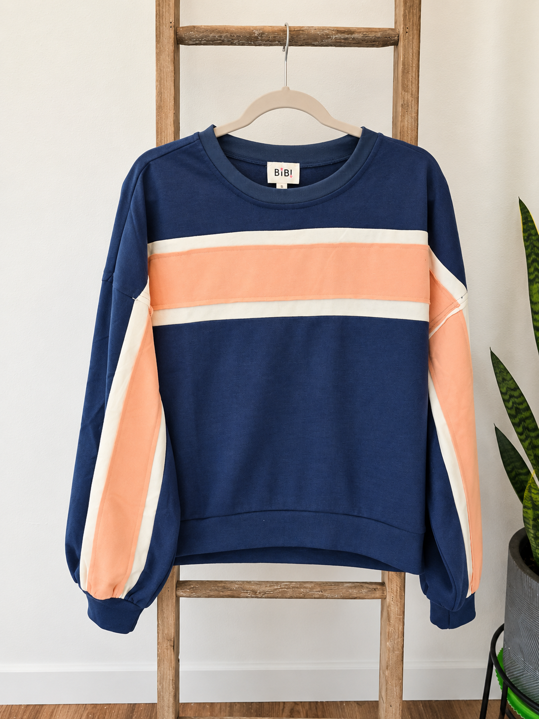 A navy blue crewneck sweatshirt with peach and cream color-blocked stripes across the chest and sleeves, hanging on a wooden hanger on a wooden ladder against a white wall.